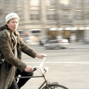 A person on a bike in a city looks into the camera, the background is motion blurred and the figure is in focus.