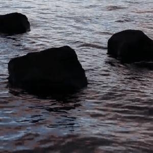 Three shadowy rocks sit up out of the bright metallic-like ocean, water swirls creating patterns around the rocks