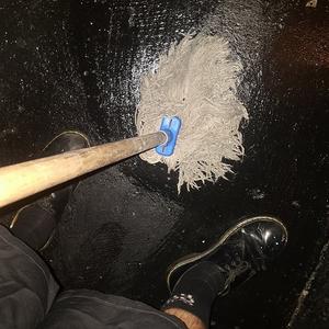 A hairy mop is being pushed against a shiny black concrete floor in a sex on site club. The image is taken looking down at the mop and the artists feet probably mid way through mopping the floor.