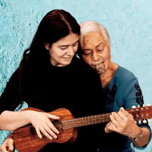 Kahurangiariki sits on their mother's lap, playing ukelele together. Behind them is a bird's eye view of water flowing