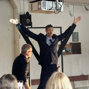 A man in a suit stands with his arms raised above his head, his arms appear to be propped up by two wooden piece sof timber.