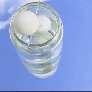 Two white ping pong balls sit inside a glass jar upon a mirror outside, the sky is reflected in the mirror and jar.