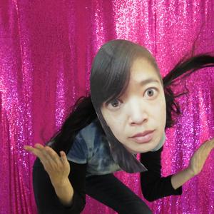 A person throws their hands up as if they are saying "what?" wearing a paper mask of Li Ming's face in front of a glitter fuschia pink curtain