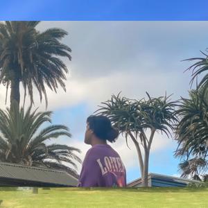 Surrounded by palm trees a person gazes into the distance, their back to the camera, the lettering on their shirt reading 'Loiter'.