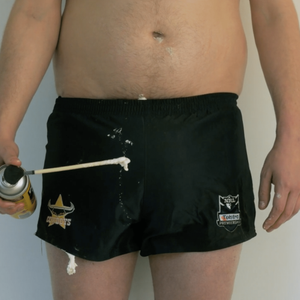 A topless man wearing short rugby shorts which are dripping with expanding foam, holds a can of expanding foam.