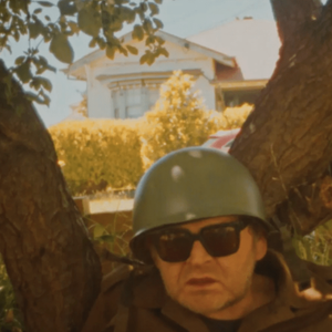 Wearing an army helmet and sunglasses, Ronnie sits against a tree with his mouth slightly open