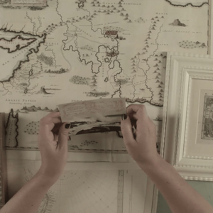 Upon a table covered with many maps, Ruth holds a postcard with two hands