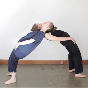 Two people stand facing away from each other 2 meters apart arching their backs dramatically in order to lean and balance upon one another