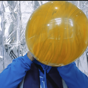 A giant yellow balloon is being blown up by a person wearing bright blue standing infront of metalic silver backrgound