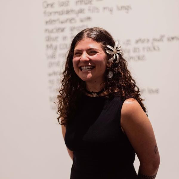 A person stands in a black dress with their arms clasped behind their back. In their hair is a white flower, they are smiling. Behind them is a blurred and obscured wall text - possibly a poem - with a header in te reo Māori.