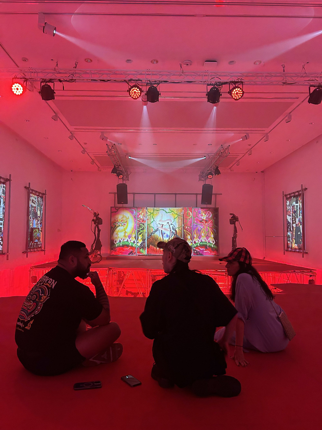 Three artists sit on a red raised platform in a gallery, in the background we see two brutalist metal sculptures. On the wall are vivid and busy collage works.