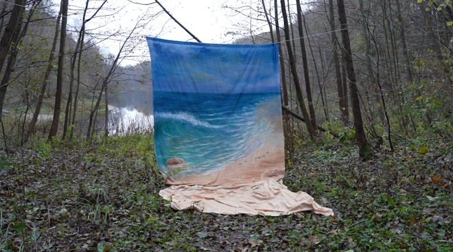 A painted fabric backdrop depicting a tropical beach is hung up between trees in a forest. A grey river can be seen in the background.