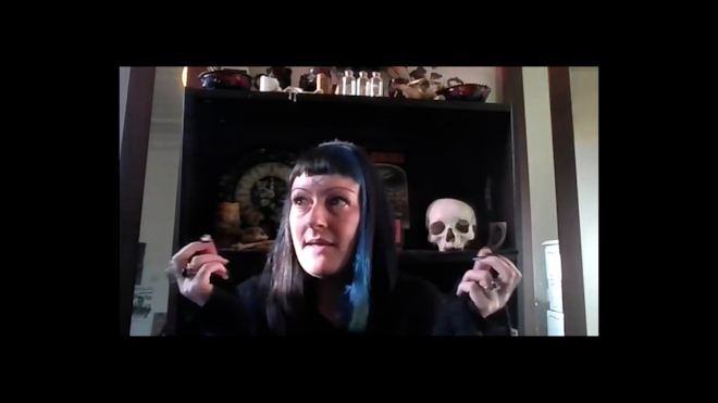 A perosn dressed in black speaks to a camera, inn the background we see a skull and occult materials.