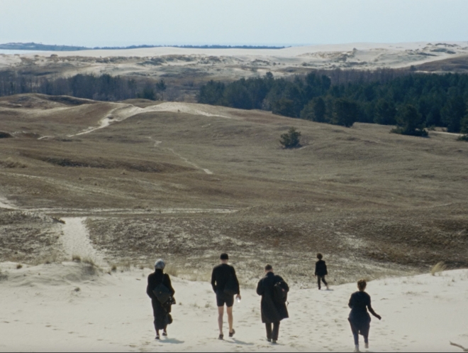 A group of people walk down a hill of sand towards a barren wilderness.