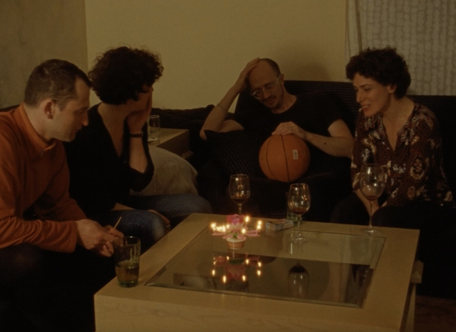 Four middle aged people are gathered around a table over drinks. One person holds a basketball in their lap. They appear to be old friends.