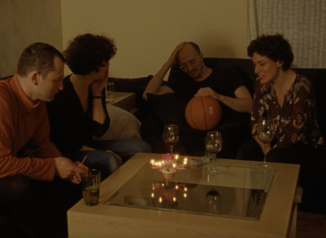 Four middle aged people are gathered around a table over drinks. One person holds a basketball in their lap. They appear to be old friends.