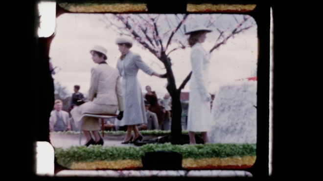An 8mm mvie still showing well dressed ladies standing and sitting by a tree. They appear to be elevated as if on a parade float. Sprocket holes for the 8mm film are visible.