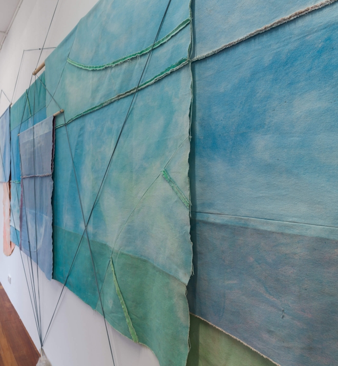 A long wall of mounted paintings, which are draped over each other. Most of the paintings share a palette of washed out blue-green and evoke the divide between land and sky.