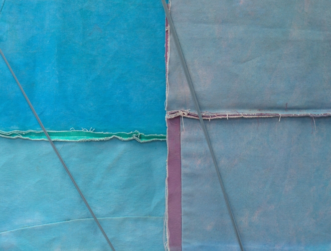 Two paintings overlap. Each is made of canvas, and has been visibly stitched together. The edge of the painting and stitching are very geometrical, evoking the lines of a flag. Two strings are tensioned across the paintings, as if anchoring something out of the picture to a point above.