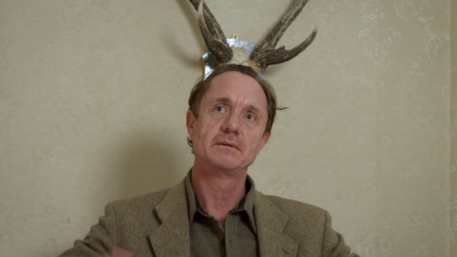 A man sits under a pair of deer antlers. He is dressed in a drab brown suit which matches the antlers.