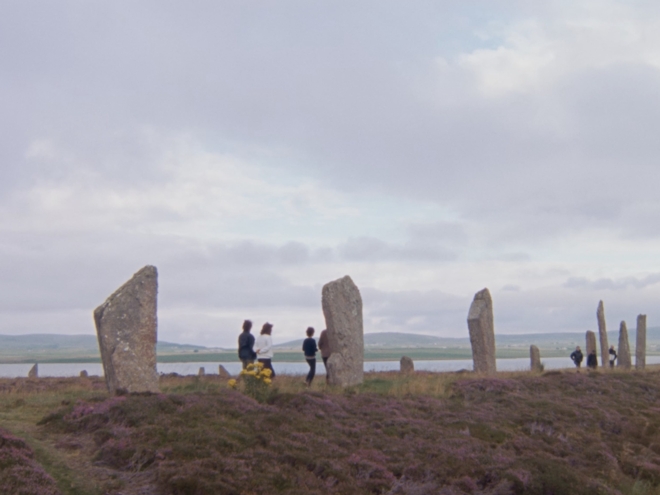 In a field by the water's edge, a group of four people walk amongst large stones. The stones are rectangular, stood on their end and are approximately 10 feet high.