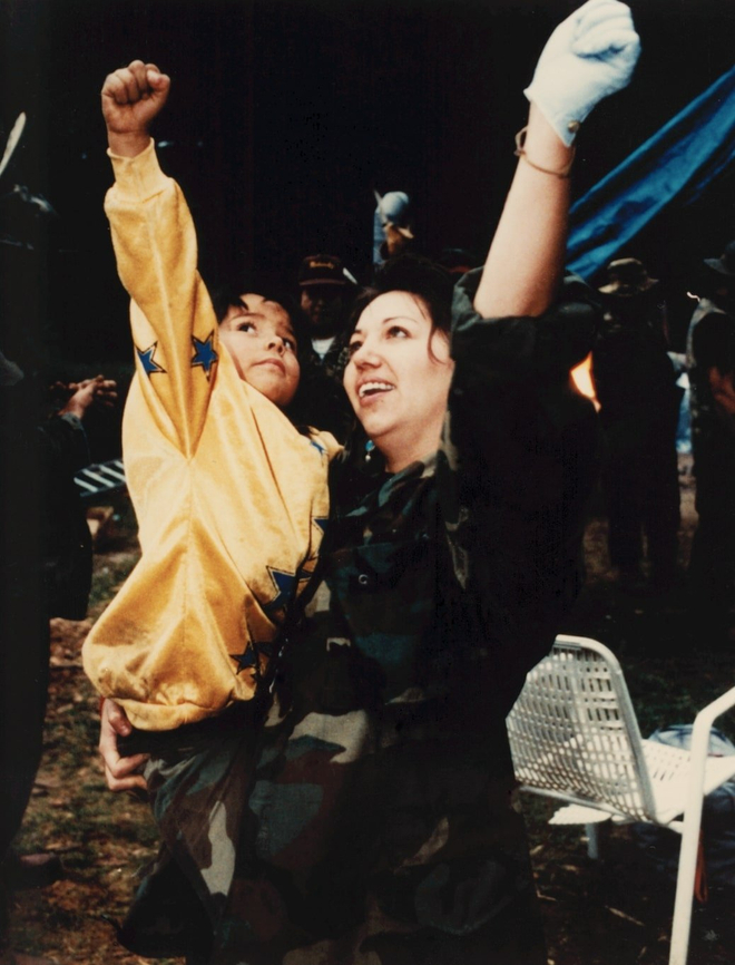 A woman carries a child on her hip; both raise a fist into the air in a gesture of resistance.