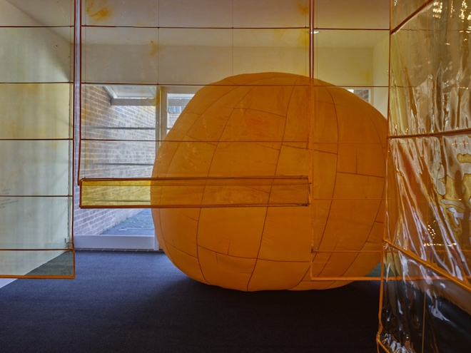 An over-sized yellow-orange sphere sits in a carpeted room. Translucent orange plastic sheets hang vertically from the ceiling in front of the ball.