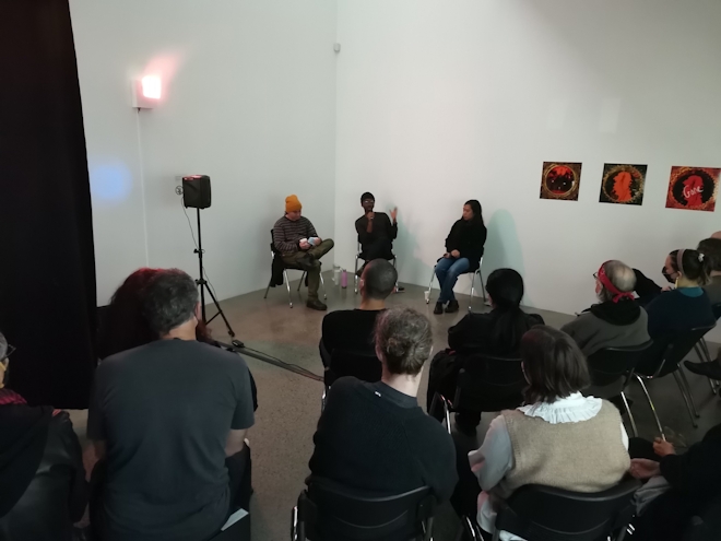 Two artists and a moderator talk in a gallery before an audience, the moderator in the centre is making a point to the audience