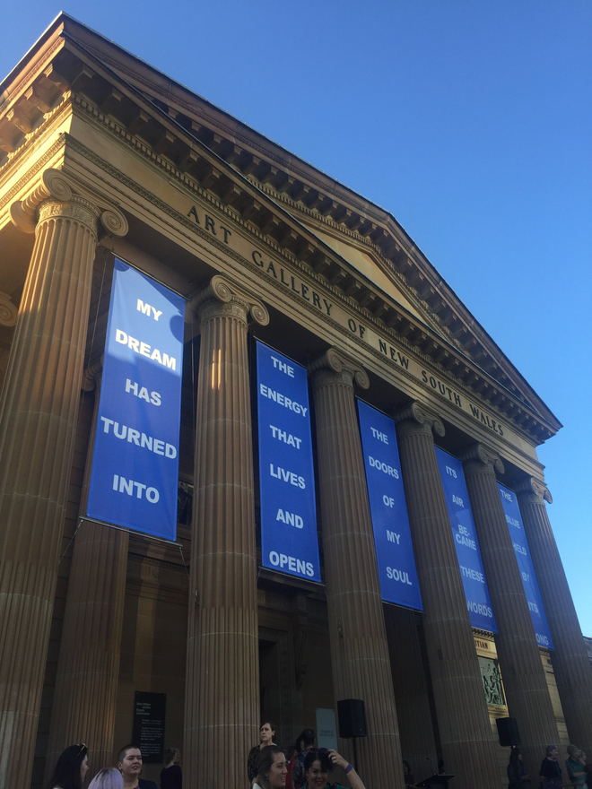 Old old stone museum building which is the Art Gallery of New South Wales has banners hanging between roman columns, the banners have text that says "my dream has turned into the energy that lives and opens the doors of my soul its air became these words he blue held by its..”