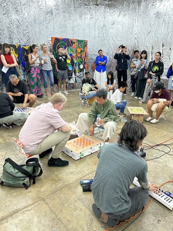 A group of people playing checkers at the opening of an exhibition while other people watch. The walls are silver foil with paintings hung on them. A musician kneels at the right foreground.
