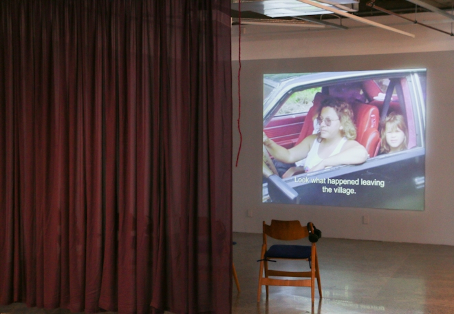 A film is projected onto a gallery wall. The still shows a woman and child in a car with the subtitle 'Look what happened leaving the village.'