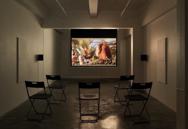 A gallery with a film showing of a person picking flowers. Empty chairs sit in the foreground awaiting viewers.