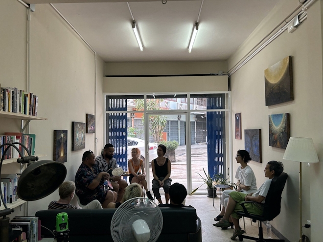 A group of people sit and meet in a circle in a gallery. Paintings are on the wall and we see the street outside.