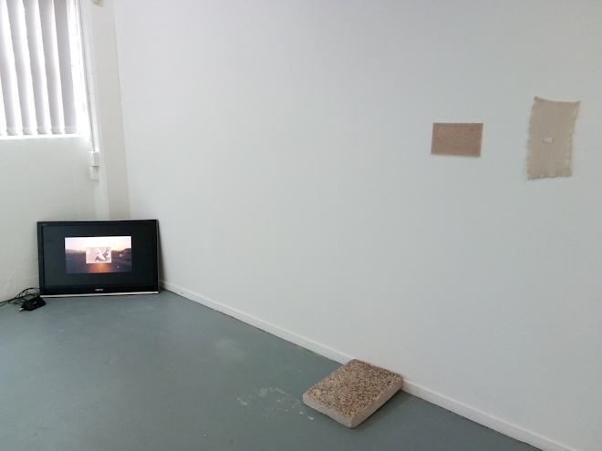 installation view of artworks made with beige stone and fabric, sittong opposite an LCD monitor showing a video work
