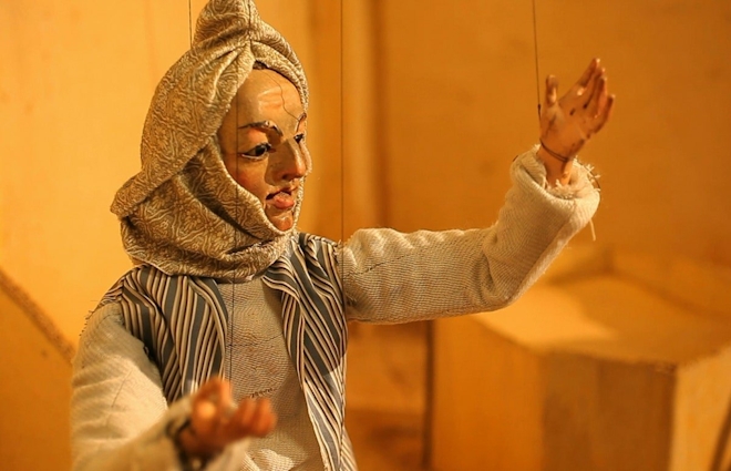 A string puppet dressed in middle eastern clothing gestures as if speaking to an audience