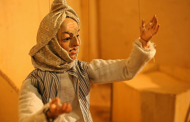 A string puppet dressed in middle eastern clothing gestures as if speaking to an audience