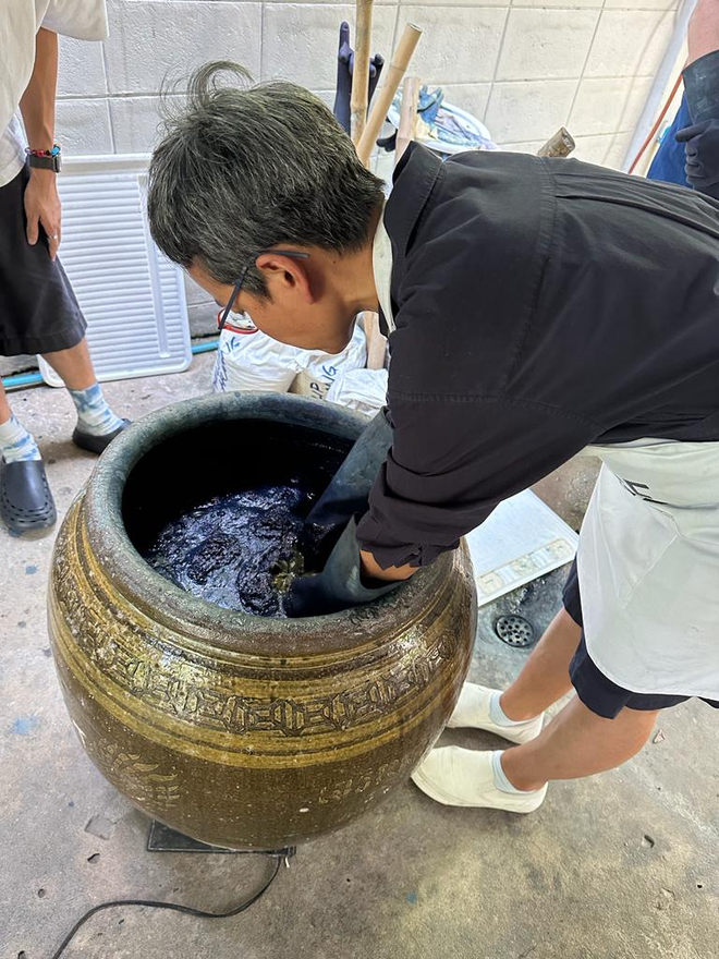 A person leans over a ceramic pot filled with purple indigo. Wearing gloves and an apron they work the fabric into the natural dye.