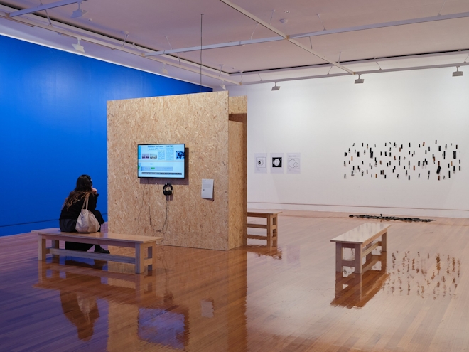 Installation shot of an exhibition showing a film work on a cork wall and various wall works. One wall is bright blue
