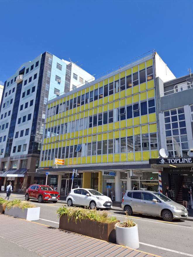 A 1960s modernist office building with yellow panelling and large windows. Cars are parked in the street below.