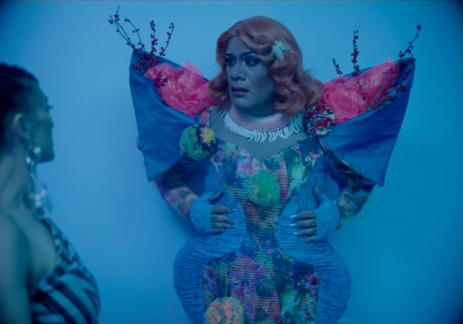 At left, a woman dressed as if for an evening at a night club encounters a woman in a floral costume augmented by exaggerated curves, pointed shoulders and foliage pointing upwards.