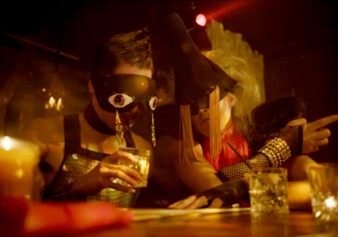 Two people in a bar drink and converse, each is dressed in elaborate masks which obscure their eyes and augment them with hanging golden adornment.