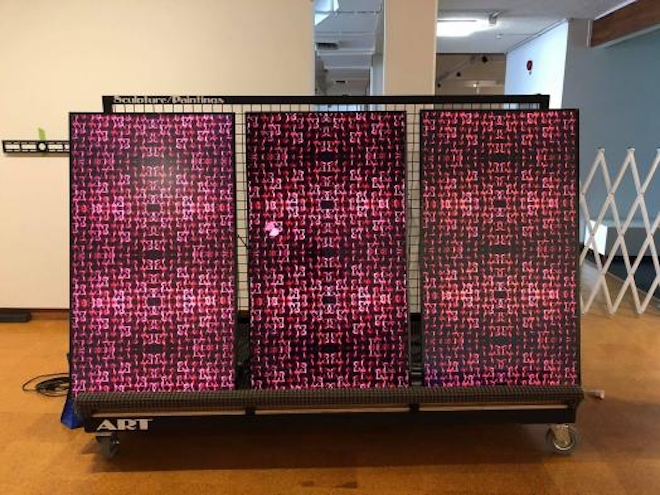 Three digital LED screens on a trolley, each showing abstract pink images that resemble abstract versions of the traditonal Māori tukutuku panel