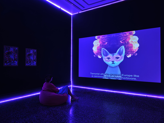 A person in a dark room is slightly illuminated by a large projection screen playing a video. The space is also illuminated by a LED lighting strip which is outlining the corners of the room. A person sits on a beanbag enjoying the video installation