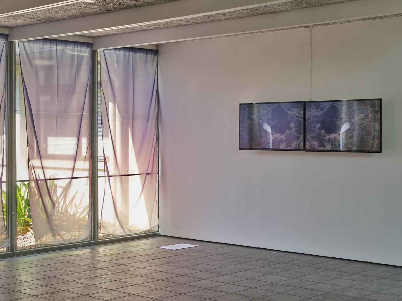 Purple-hued sheer curtains cover the windows of an exhibition space; on the wall are two video screens depicted smoke plumes from an industrial chimney.