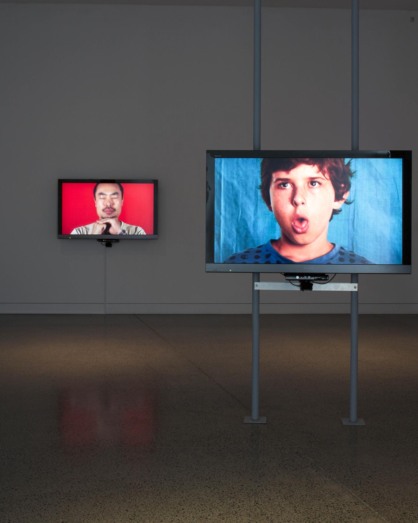 Two LCD monitors mounted in a gallery, each showing an individual making a sound with their body