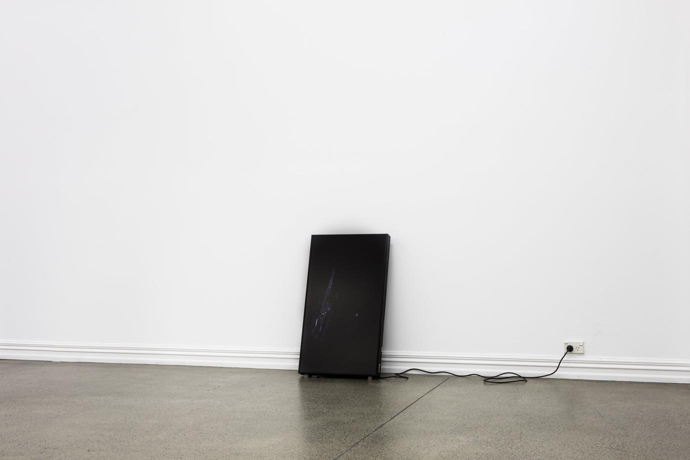 An LCD monitor with an almost imperceptably dark image stands against a wall, abjectly supported by a power cord plugged into the wall