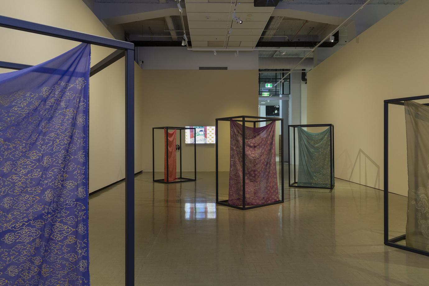 A series of colourful fabrics hang from large black rectangular frames. On the rear wall is the glow of a video monitor.