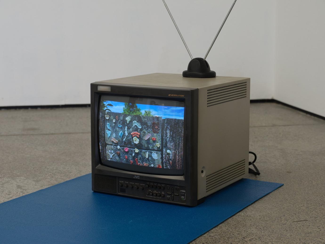 A CRT TV sits on a blue mat in a gallery space. The display depicts a colourful assortment of chewing gum affixed to a concrete wall