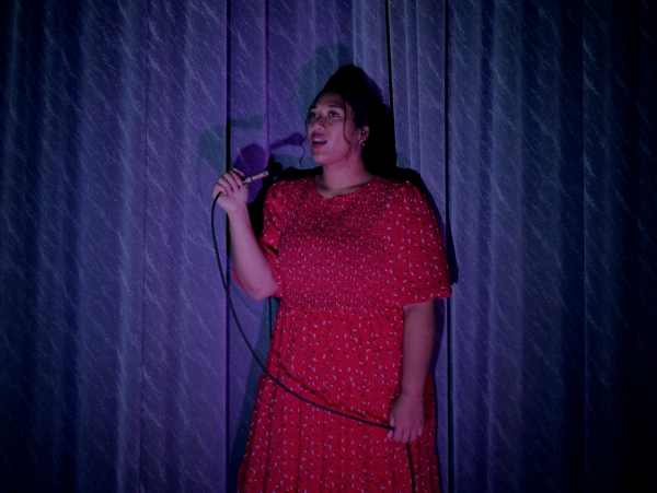 A Pasifika woman in a red dress sings alone into a microphone  in front of a blue stage curtain