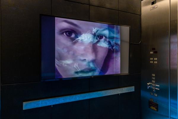 Inside a lift, screen showing Kate Moss's face with animated figures overlaying it.